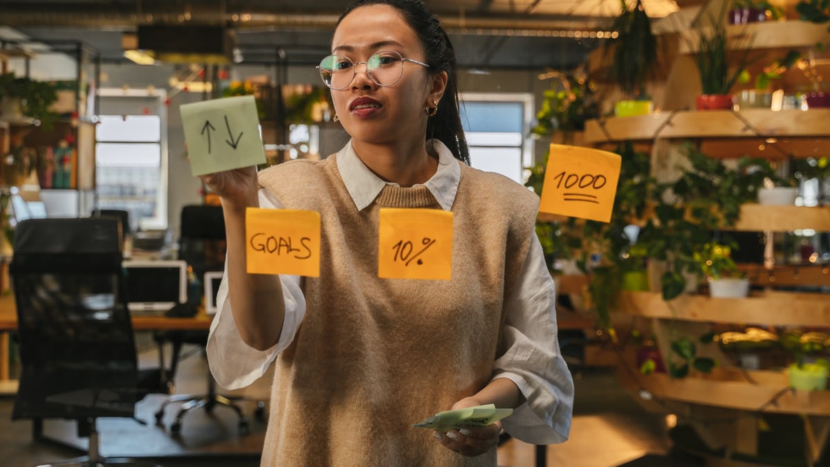 Asian woman organizing tasks on sticky notes at modern office workspace.